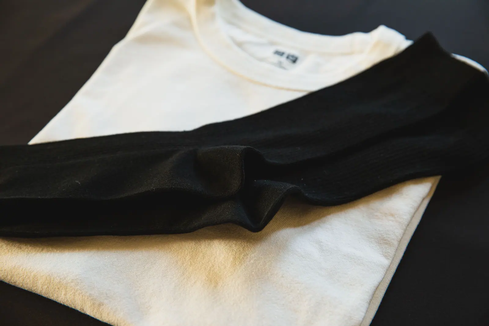 A folded cream T-shirt with a black sock laid across it on a dark surface.